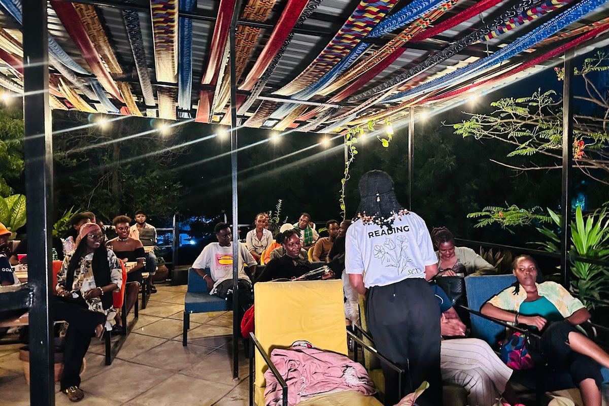 An evening gathering under a canopy of hanging printed fabric strips, with the audience seated in armchairs and a speaker standing.