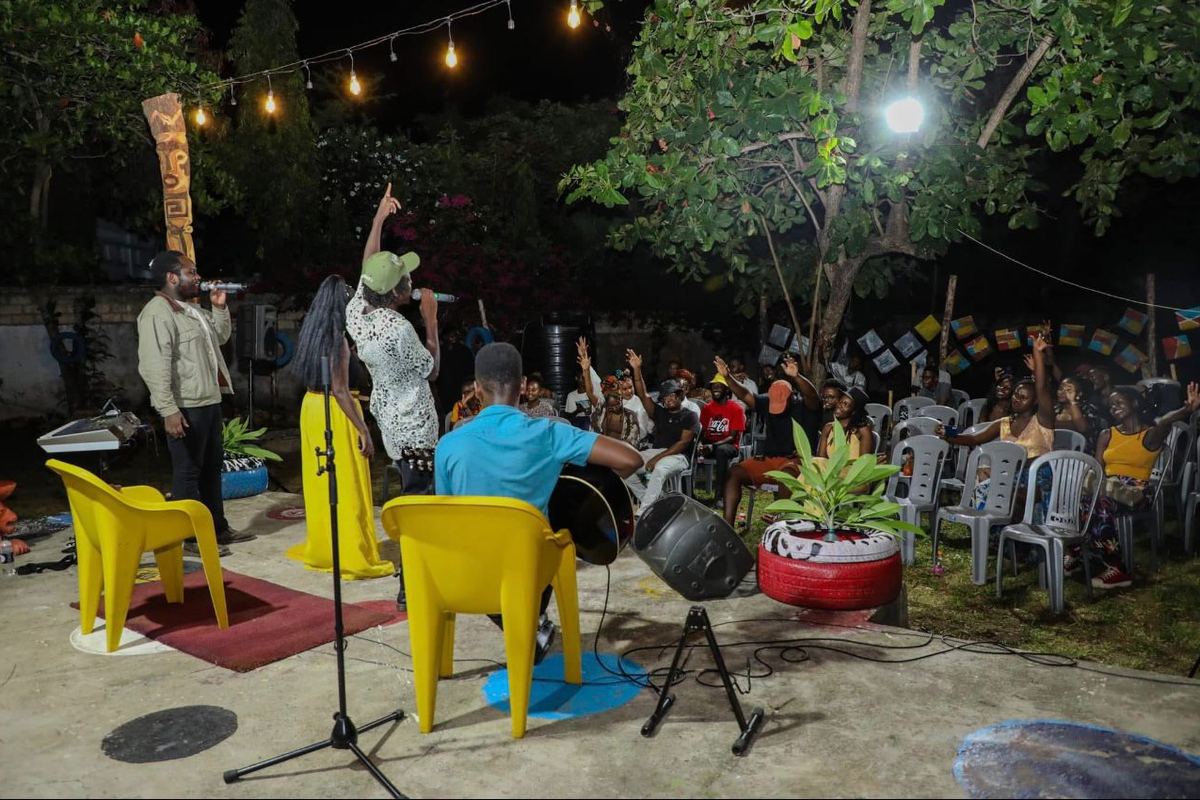 An outdoor evening performance at Africa Nomads Art Space, two singers on a lit concrete stage with a seated audience raising their hands.