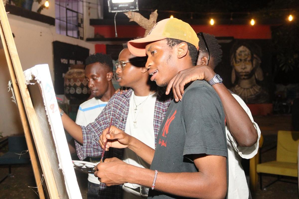 Four young men painting together at a single easel during an evening session, arms around each other's shoulders.