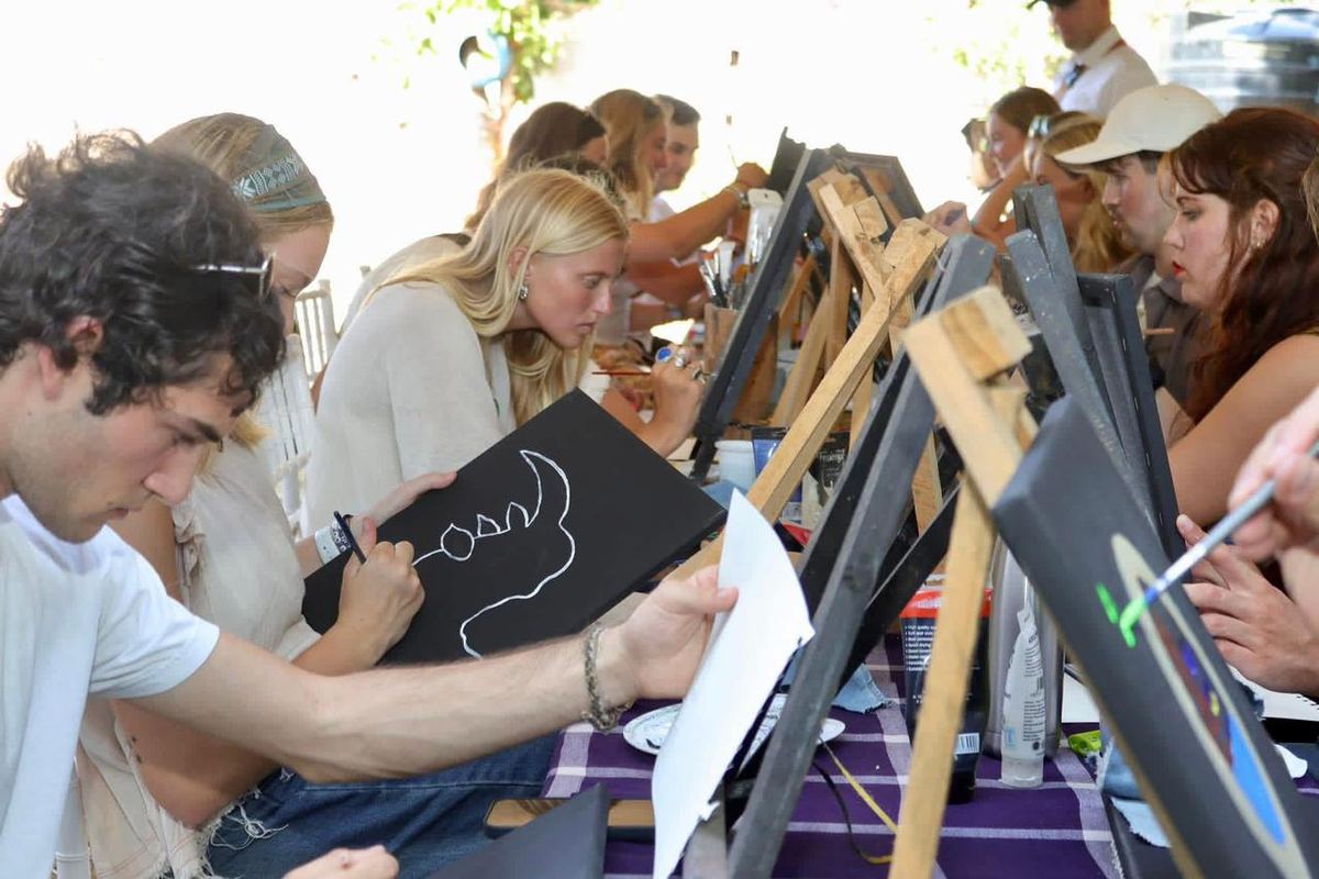 Participants at a long table during a painting workshop, each working on a small canvas on an easel.